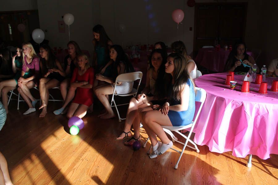 Group of young people seated in a room with a pink table, balloons, and sunlit wood floor.
