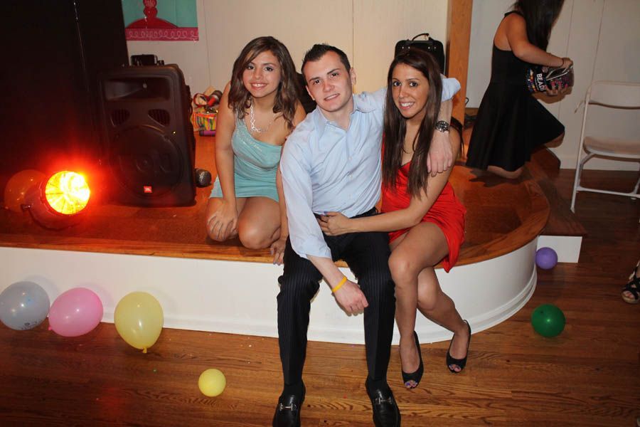 Three people pose on a stage with balloons. A man sits between two women, all smiling.