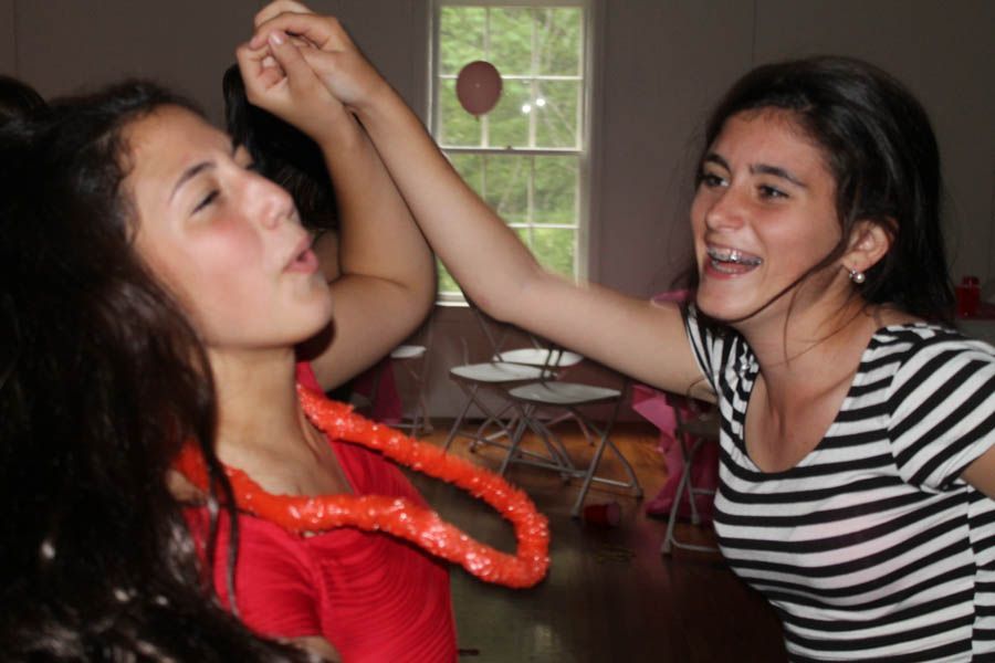 Two girls laughing and playing with a lei indoors; one wears a striped shirt, the other a red top.