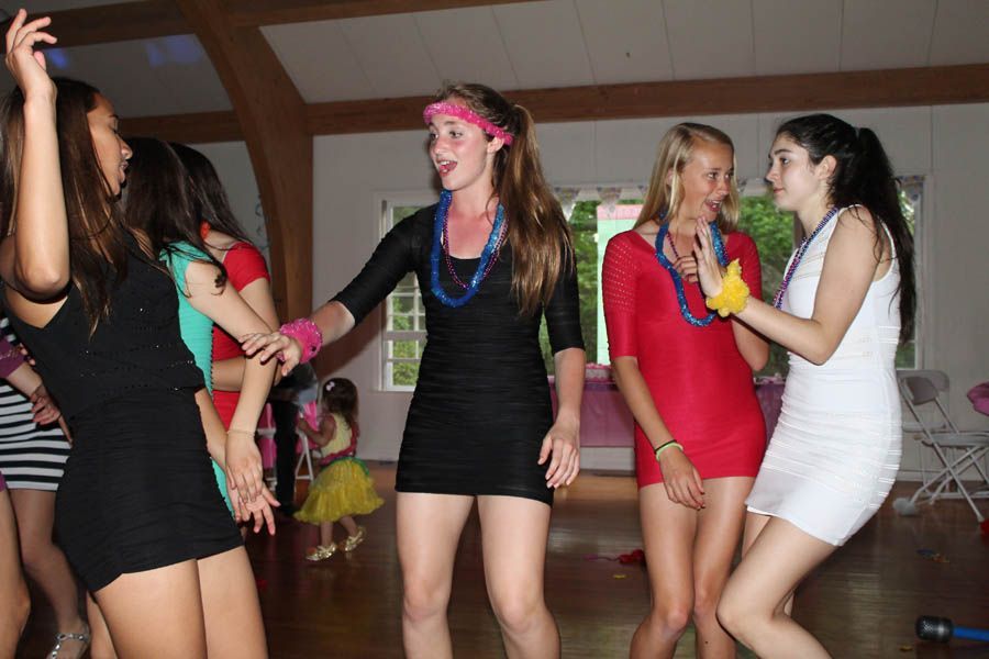 Group of people dancing indoors, wearing short dresses, some with accessories, bright colors.