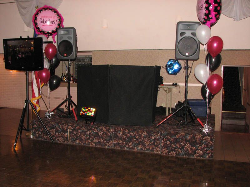 DJ setup with speakers, screen, and black booth on a decorated stage; balloons in dark pink, white, and black.