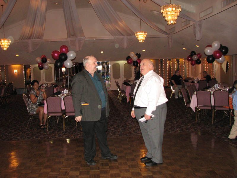 Two men talking on a dance floor in a decorated ballroom with balloons, tables, and chandeliers.