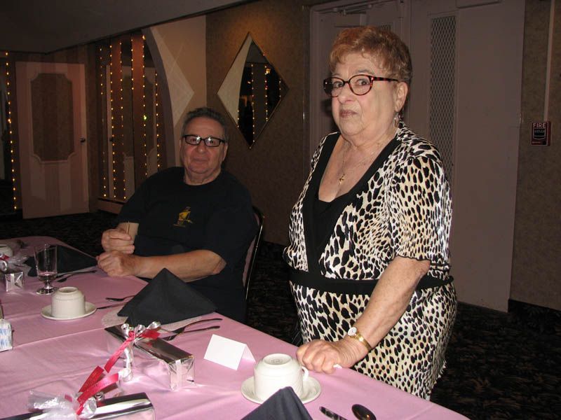 Two people at a table; woman in animal print dress, man in black shirt, pink tablecloth, and glasses.