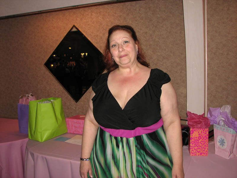 Woman in black top and green dress, standing at a table with gifts. Interior setting with decorative wall.