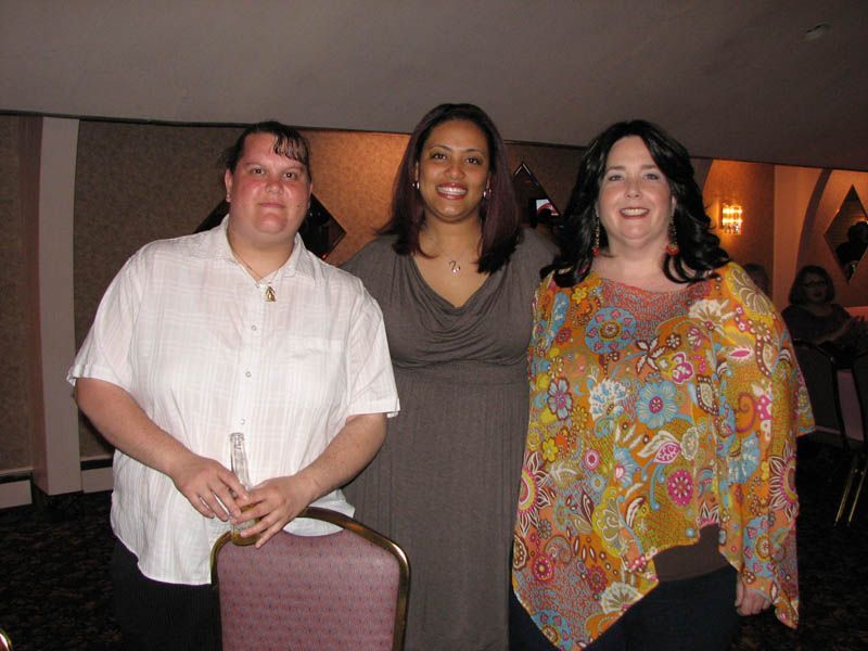 Three women posing indoors at an event. The woman in the center wears a brown dress; the others wear tops.