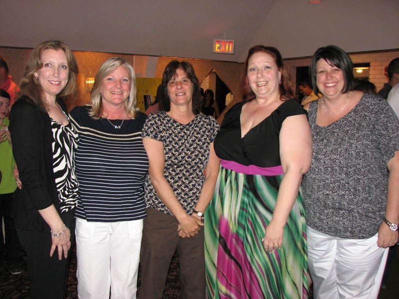 Five women smiling, posing for photo indoors. One in black and white print, others in casual attire.