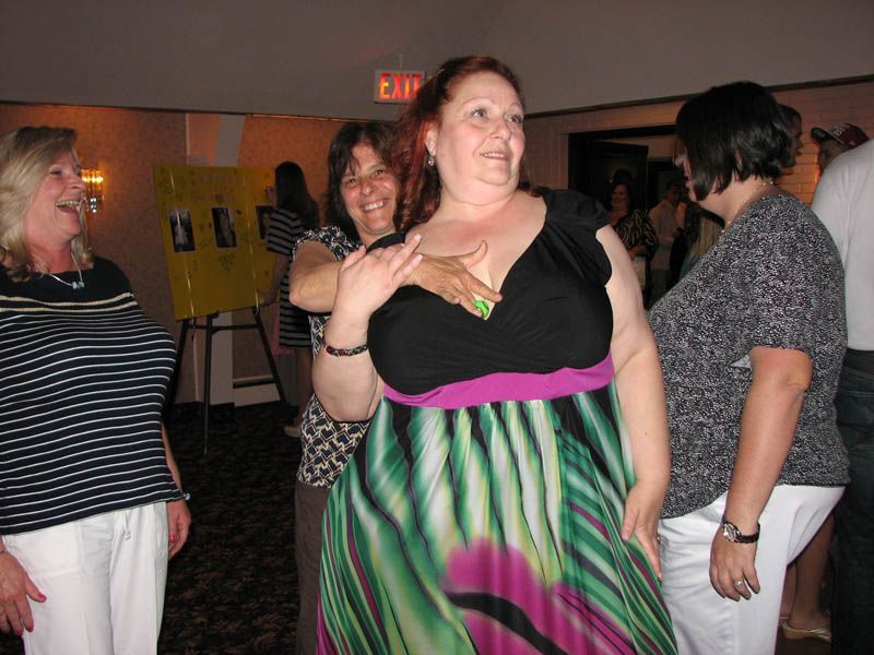 Four women at an event; woman in center in a colorful dress smiles as another touches her chest.