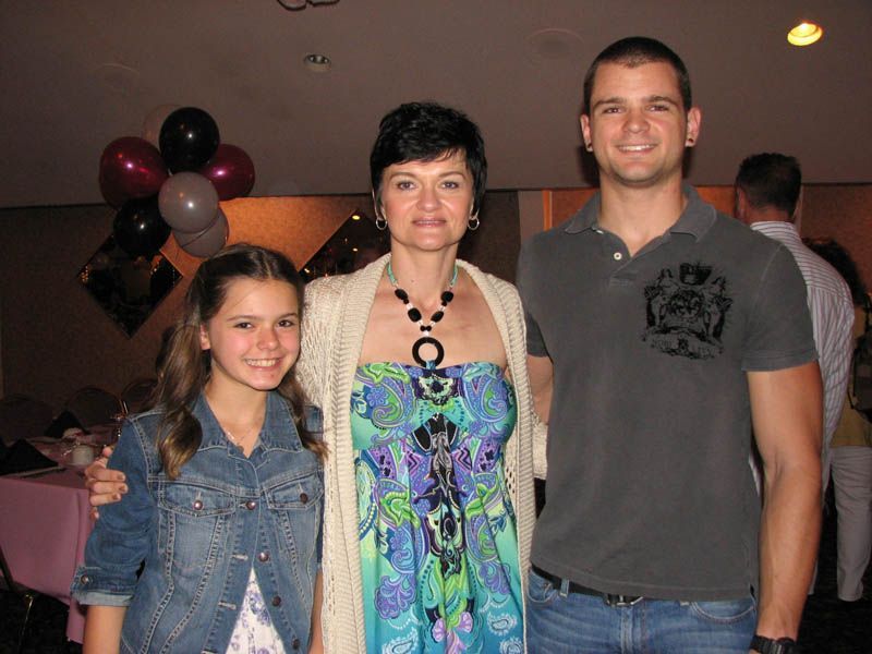 Woman with two younger people, arm around each other, near balloons and tables.