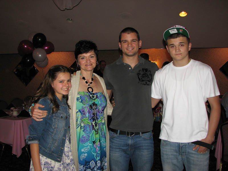 Family posing together indoors at an event. A woman, two young adults, and a girl smiling. Balloons in the background.
