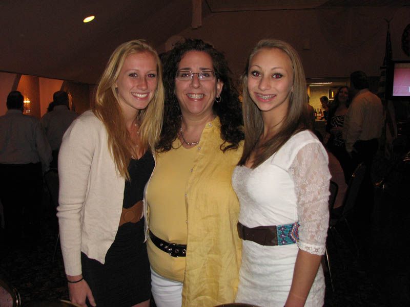 Three people pose for photo. One in cream sweater, one in yellow top and scarf, one in white dress. Interior setting.