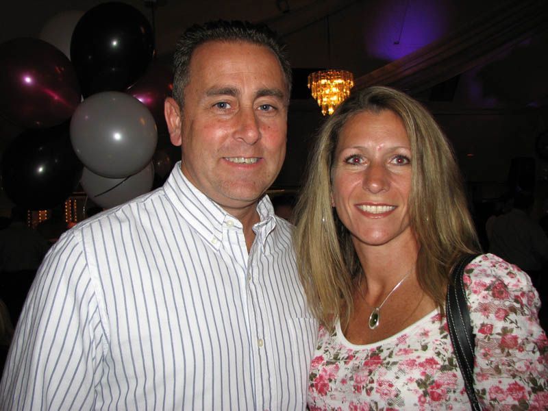Man and woman smiling together, indoors near balloons, the woman with long blond hair and a floral shirt.