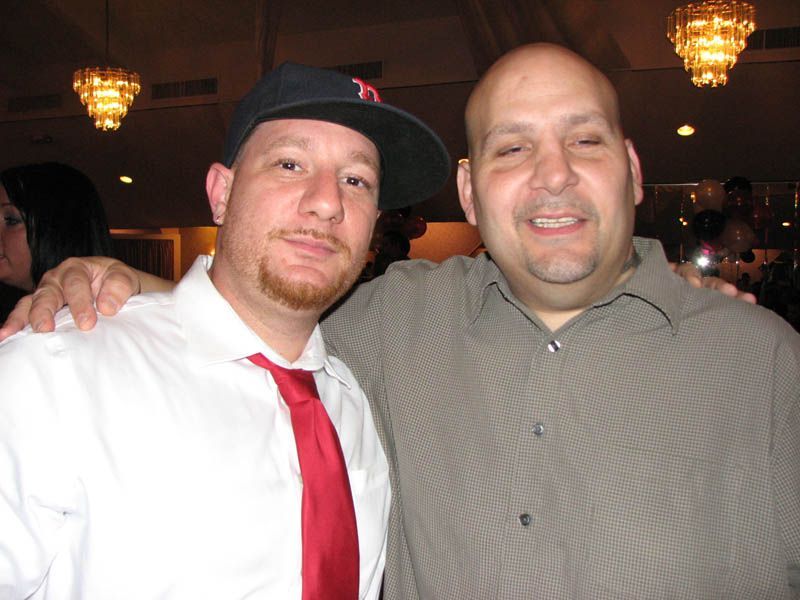 Two men smiling, one in a cap and tie, the other with hand on the cap's shoulder. Indoors, chandeliers.