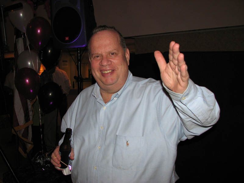 Man in light blue shirt waves, holding a bottle, smiles at a party with balloons.