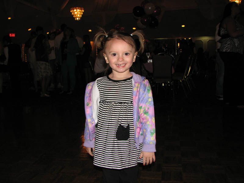 Young girl with pigtails smiles, wearing a striped dress and floral sweater, in a dimly lit event space.
