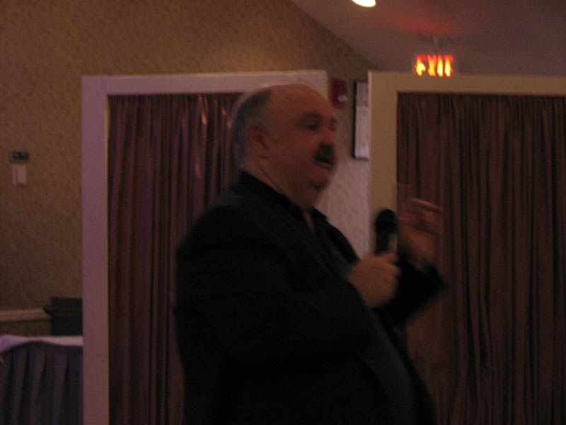 Man speaking into a microphone, gesturing with his hand in a room with burgundy curtains.