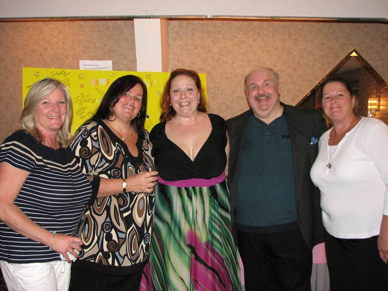 Five people standing together, smiling. Women in patterned and solid-colored tops. Man in blazer. Party setting.