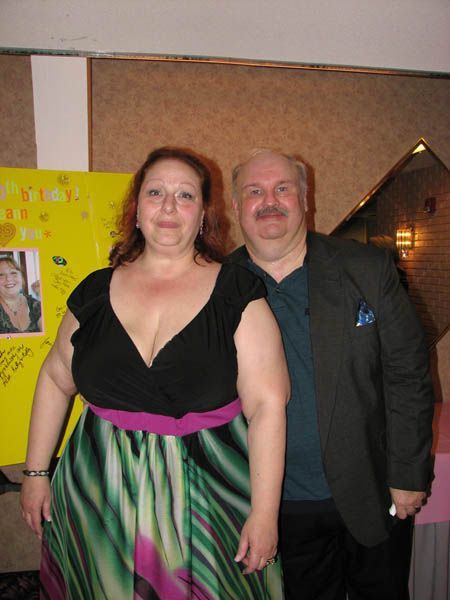 Woman in black top and colorful dress, man in jacket pose near a poster.