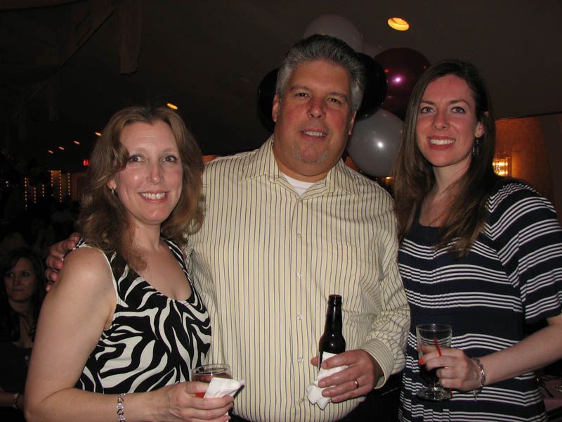 Three people smiling at a party. The man holds a beer bottle, the women hold drinks. Balloons in the background.