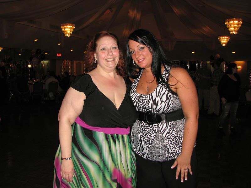 Two women smiling indoors. One wears a black top, colorful skirt; the other a zebra-print top. Dim lighting.