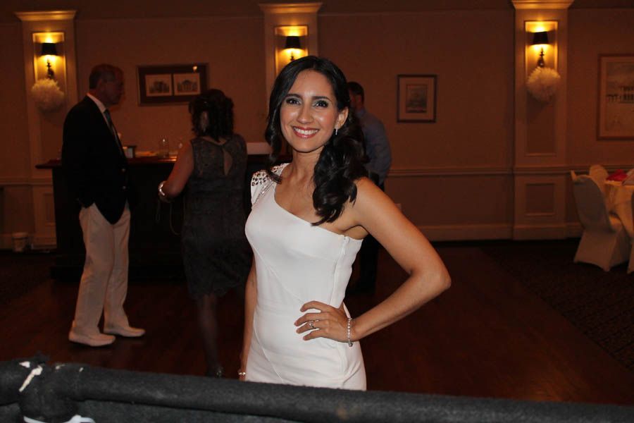 Woman in white dress smiles, poses at an event. Dark hair, hand on hip, warm-toned room.