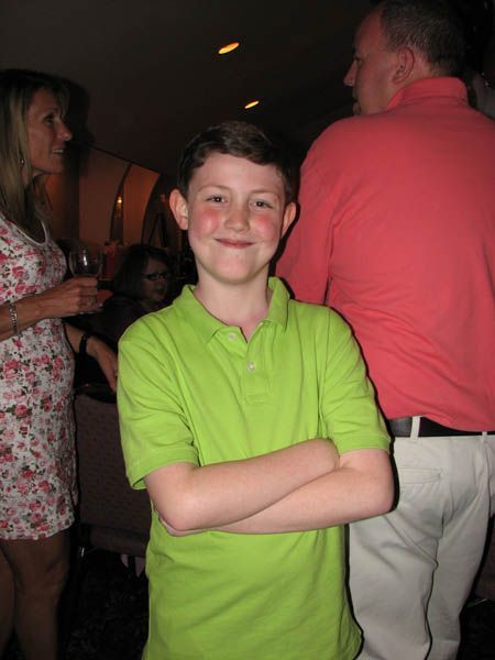 Boy in green polo shirt, arms crossed, smiling in a room. People in background.