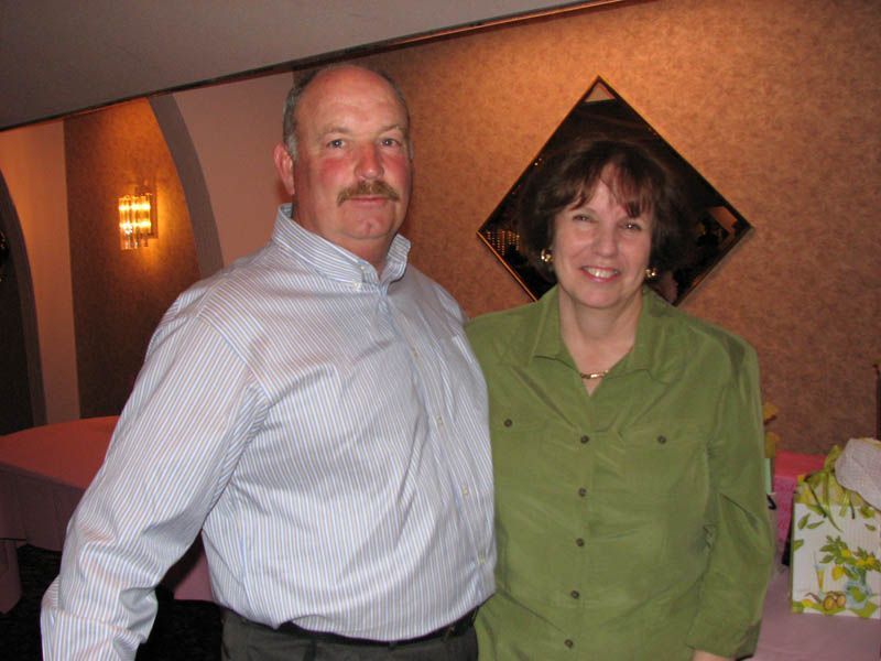 Man with mustache in blue shirt stands beside a woman in green shirt, posing together indoors.