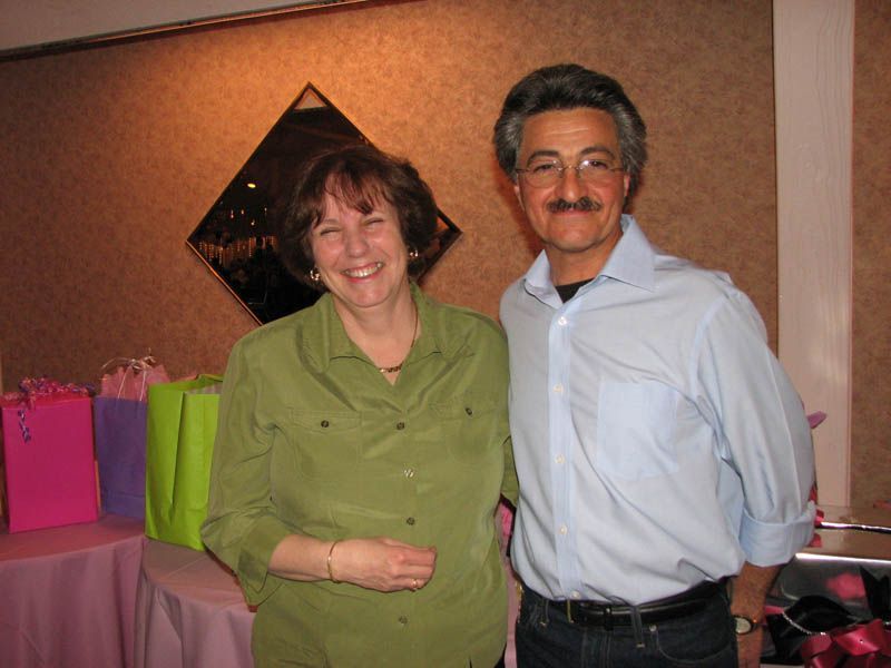Two smiling people stand in front of a table with gift bags. The woman wears a green shirt, and the man wears a blue shirt.