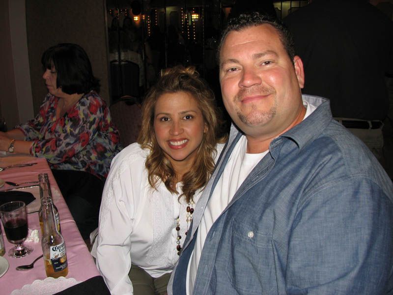 Woman and man smiling, posing indoors. Woman in white shirt, man in denim. Table setting in foreground.
