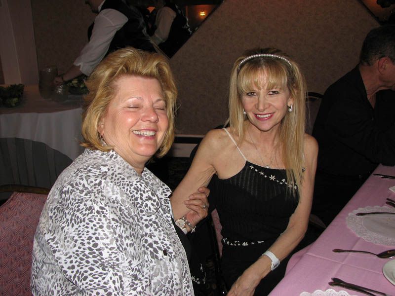 Two smiling women at a table; one wearing a black dress, the other a patterned jacket.
