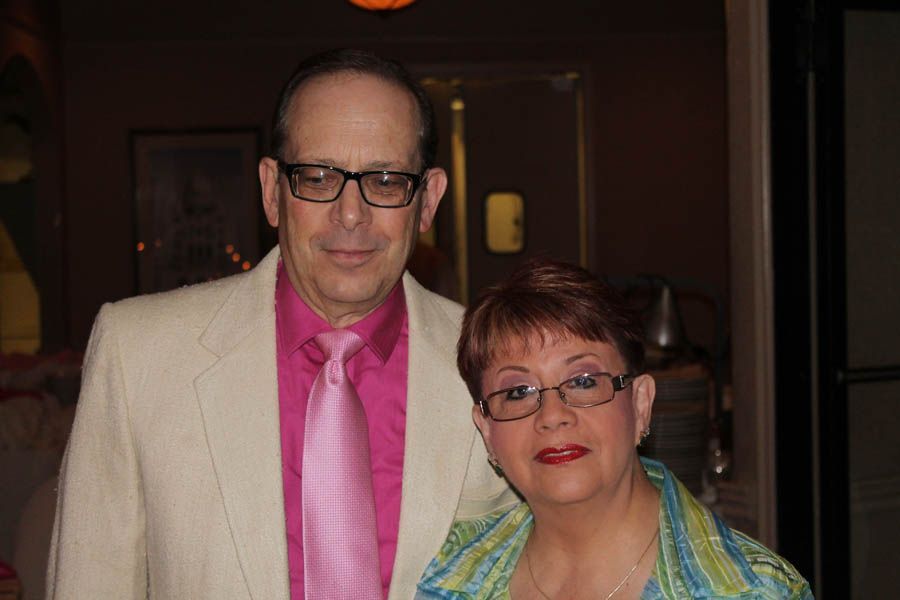 Man in light suit, pink shirt/tie, and woman in glasses stand side-by-side, smiling indoors.