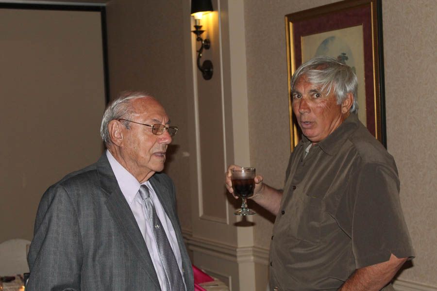 Two men talking in a room, one holding a drink. The setting is indoors, with tan walls and a framed artwork.