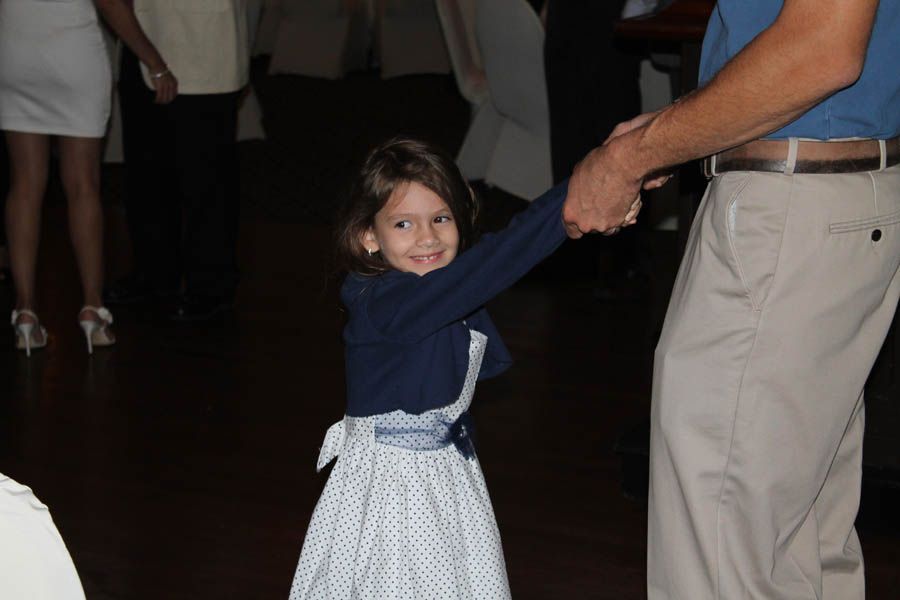Girl dancing with a man, holding hands. She wears a dress and jacket, smiling. Dark room, other people visible.