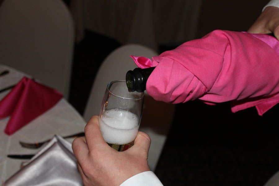 Person pouring champagne into a glass. Bottle wrapped in pink cloth; table setting in background.