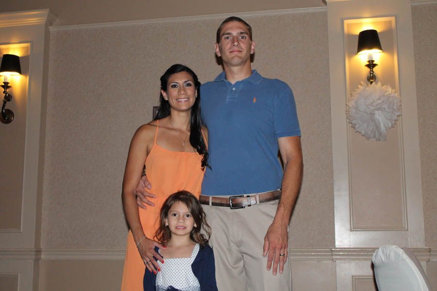 Family of three smiles for photo. Woman in orange dress, man in blue shirt, child in blue cardigan.