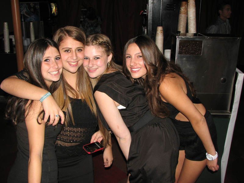 Four young people, smiling and hugging, in a dimly lit indoor setting. All are wearing black dresses.