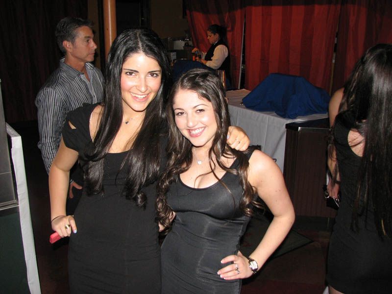 Two women in black dresses pose together, smiling at a party.