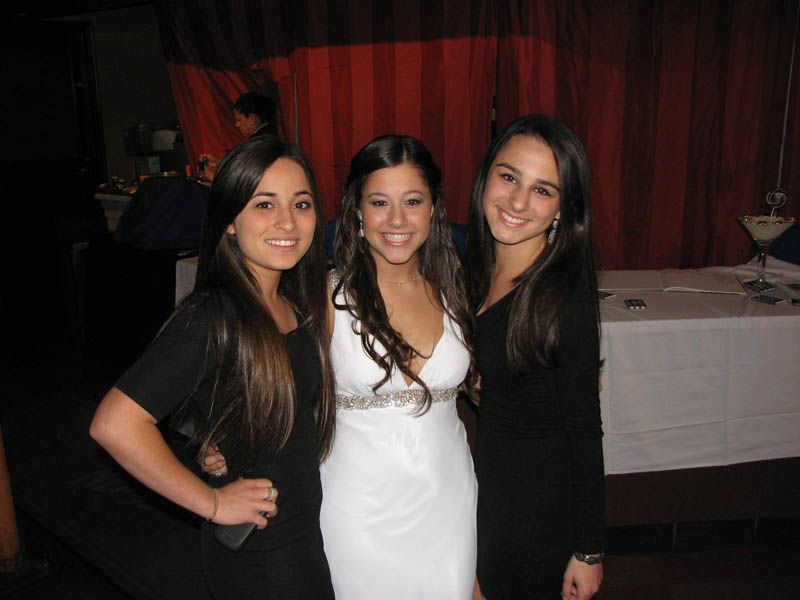 Three women smiling, posing for a photo. One in white gown, others in black dresses, indoors with red curtains.