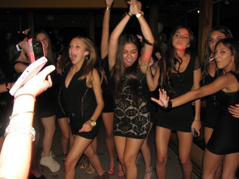 Group of women dancing at a party; most in black dresses, arms raised.