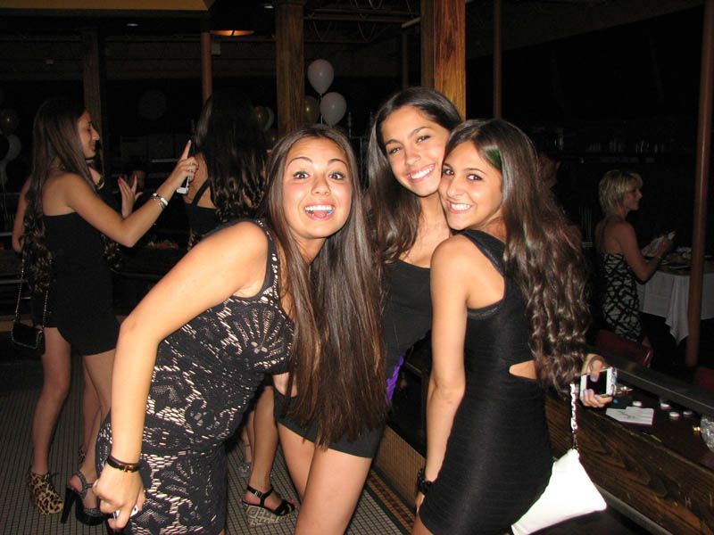 Three young women smiling, posing at party; one in black lace dress, two in black dresses.