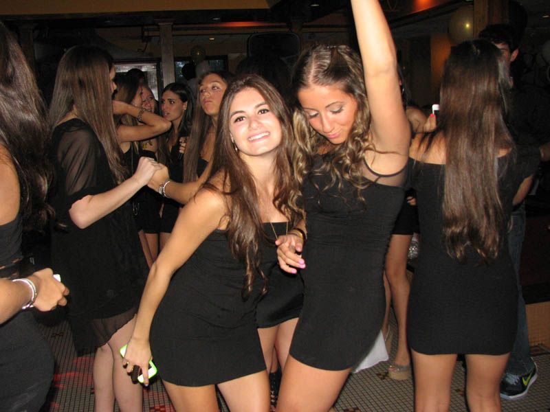 Group of women dancing at a party in black dresses, smiling and raising arms. Dim lighting.