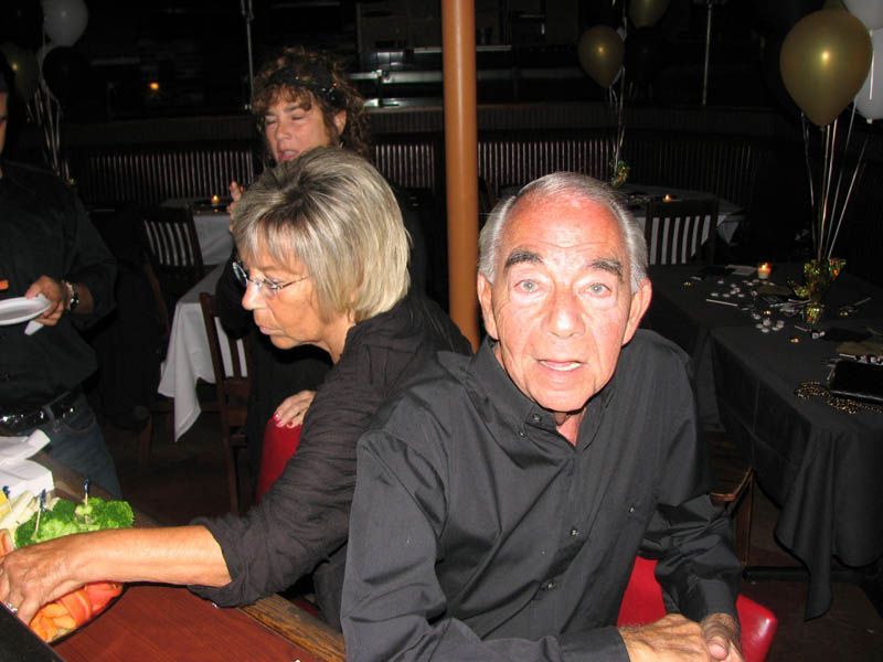 Older man in a black shirt at a party. Other people, balloons, and tables are in the background.