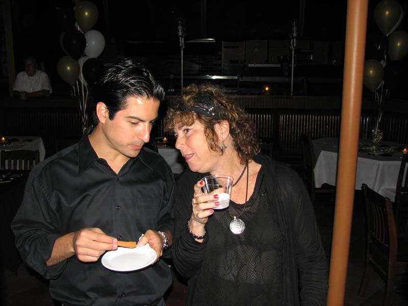 Man and woman in dark clothing, at an event. Man holds a snack on a small plate. Woman holds a drink.