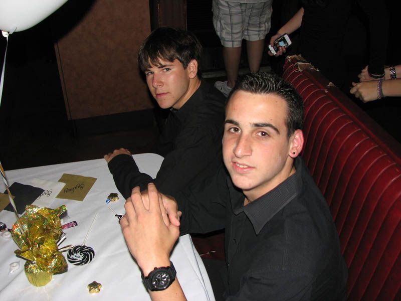 Two young men seated at a table in a dimly lit room, both wearing black shirts. One looks at the camera.