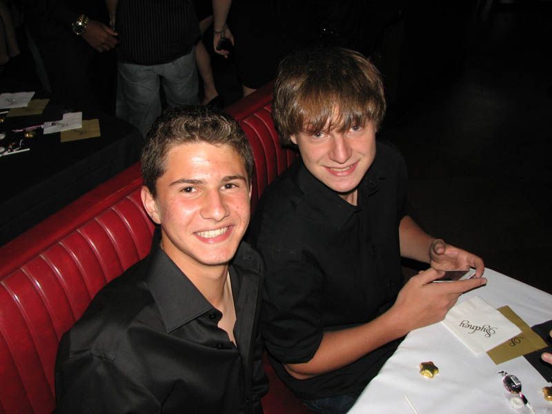 Two young men smiling at a table with a red booth. They wear black shirts, likely in a dark venue.