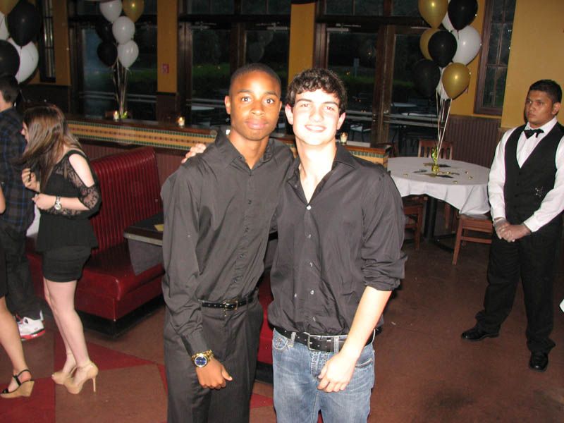 Two young men in black shirts pose for a photo at a formal event, with balloons and a waiter in the background.
