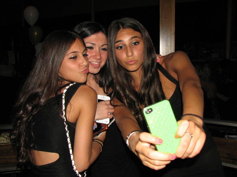 Three people in black dresses take a selfie; one is holding a green phone, the others pout.