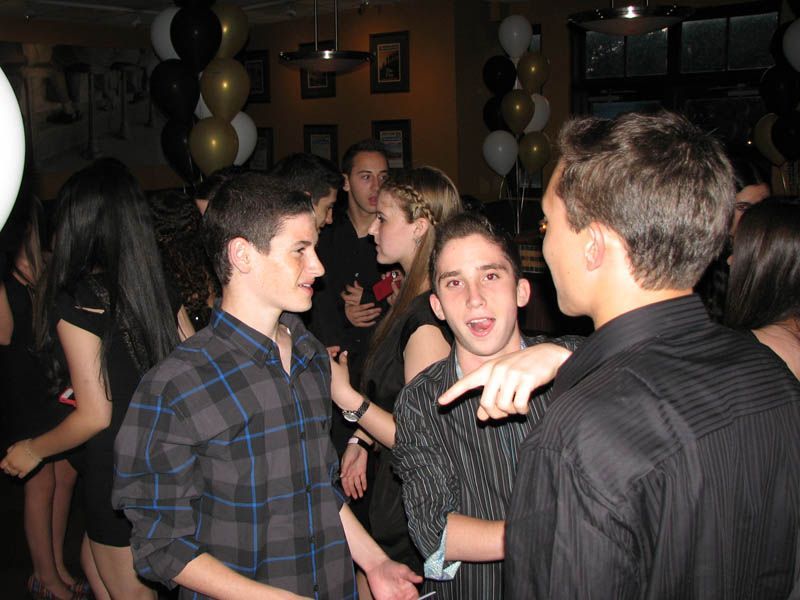 People at a party, mostly young adults, conversing in a dimly lit room with black and gold balloons.