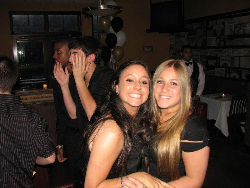 Two smiling women pose at a party. Other people are in the background, one with hands up. Balloons are visible.