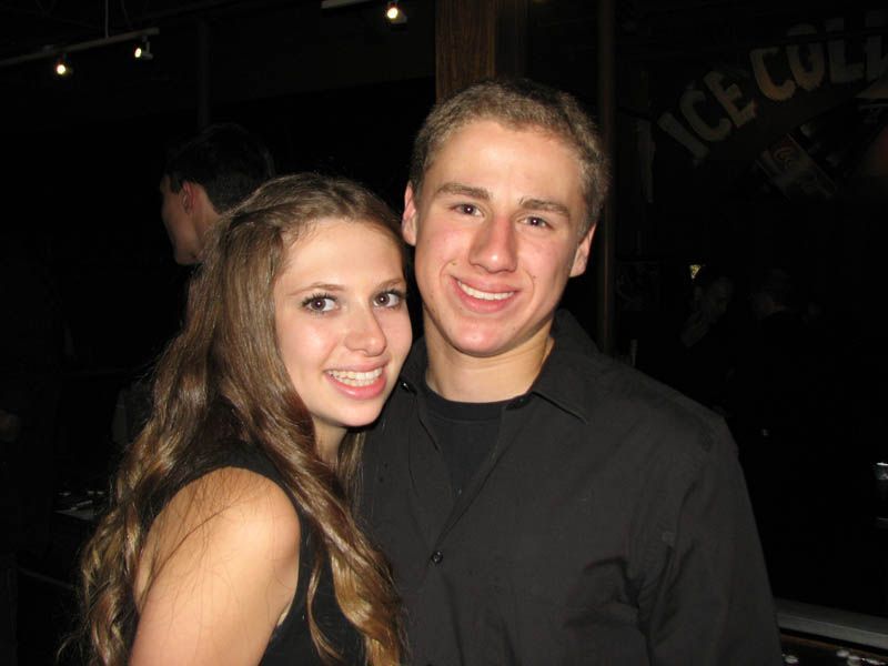 Young couple smiles, posing close together indoors; man wears black shirt, woman wears a dark sleeveless top.