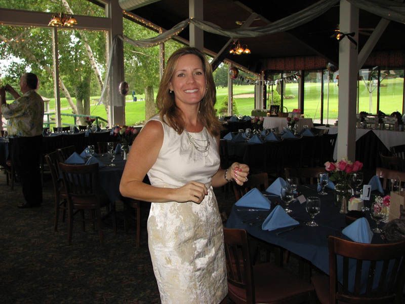 Woman in a cream dress smiles indoors, surrounded by tables set with blue tablecloths and napkins.